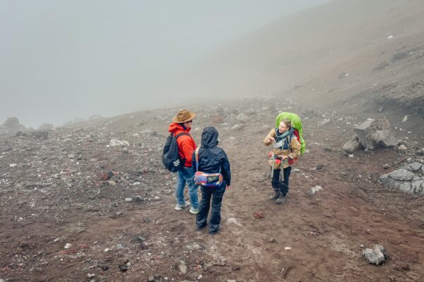 Hikers in colorful jackets stand on rocky terrain at the Jose Rivas Refuge, surrounded by fog on Mount Cotopaxi, Ecuador.