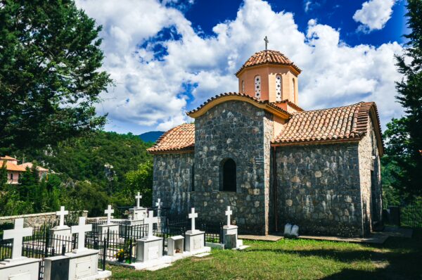 Panagia Kernitsis Monastery in Arcadia features stone architecture, a tiled roof, and a nearby graveyard under a bright blue sky.