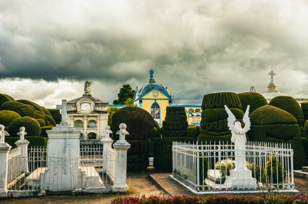 Well-manicured hedges and ornate white statues surround colorful mausoleums at Tulcán Cemetery, Ecuador, under a dramatic sky.