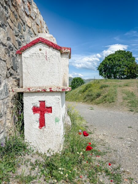A small white chapel marked with a red cross stands beside a gravel path in Vytina, Arcadia, Greece, surrounded by greenery.