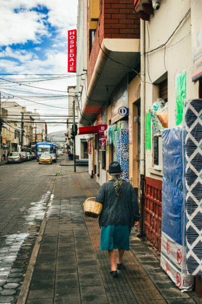 A woman carrying a basket walks along a colorful street in Ibarra, Ecuador, with shops and blue skies visible.