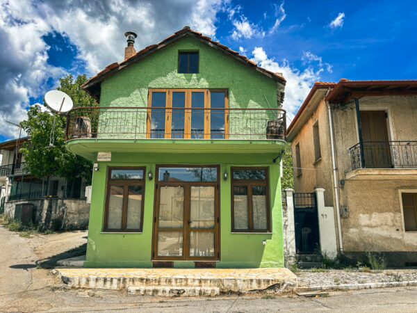 Bright green house with large windows and balcony in Nymfasia, Arcadia, Greece, framed by scattered clouds and neighboring buildings.