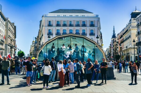 Crowd gathers around a vivid art installation in Puerta del Sol, Madrid, showcasing lively scenes under a clear blue sky.