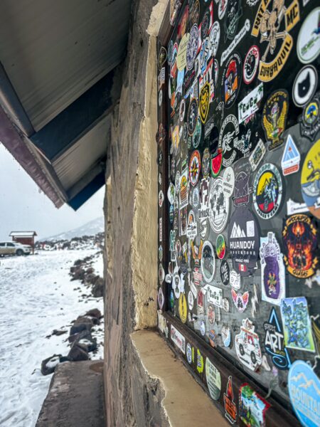Colorful stickers cover a window at Refugio Carrel, with snow-covered ground and Chimborazo Volcano in Ecuador visible nearby.