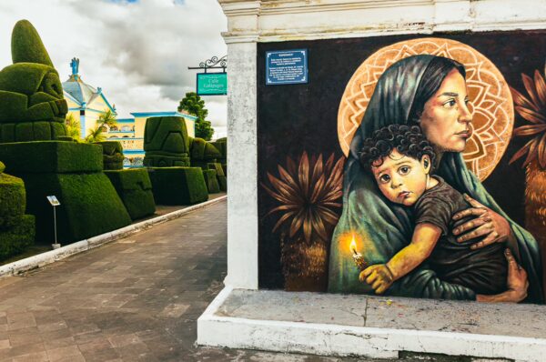 Murals of a woman and child contrast with lush green topiary at Tulcán Cemetery in Ecuador, showcasing vibrant colors and artistry.