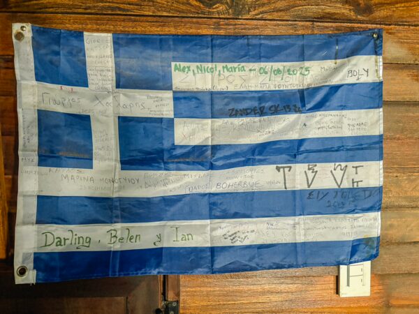 A Greek flag hangs against a wooden backdrop, featuring blue and white stripes with a prominent cross and various signatures.