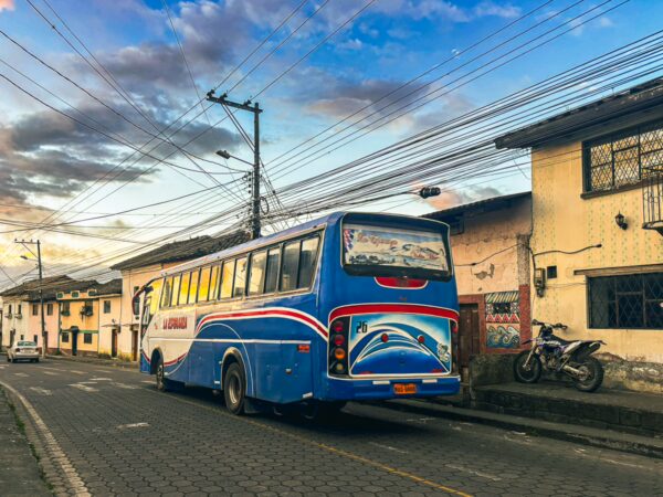 Bright blue bus parked on a street in La Esperanza, Ibarra, Ecuador, with colorful buildings and a dramatic sky in the background.