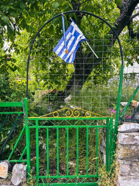 Blue and white Greek flag hangs above a green garden gate, surrounded by leafy foliage in Nymfasia, Arcadia, Greece.