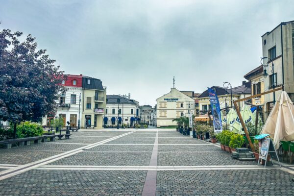 Cobblestone square in the Old Town of Craiova, Romania, lined with colorful buildings and greenery under a cloudy sky.