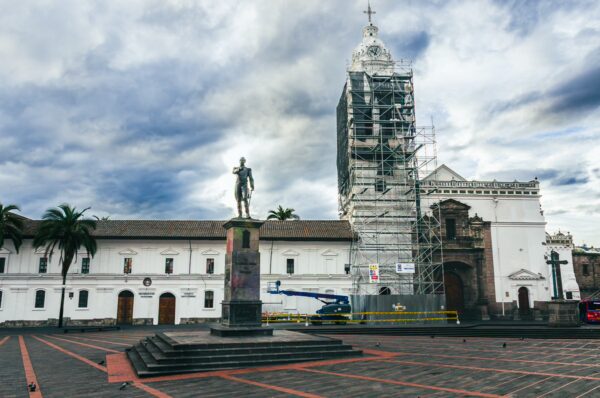 Statue in Plaza de Santo Domingo with the Cathedral in restoration, surrounded by palm trees under a cloudy sky in Quito, Ecuador.