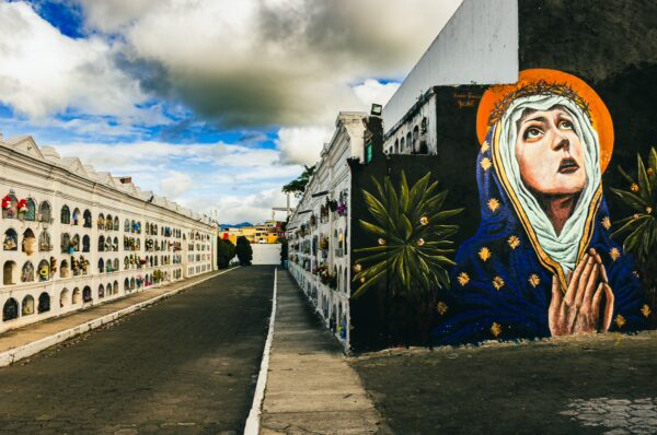 Colorful mural of a woman in prayer alongside the white tombs at Tulcán Cemetery, Ecuador, under a partly cloudy sky.