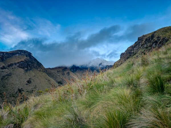 Lush green grasses rise on the slopes of Imbabura Volcano, with clouds partially obscuring its peak against a bright blue sky.