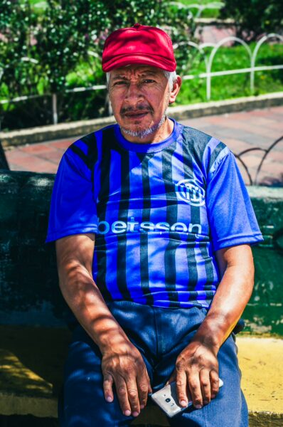 A man in a blue and black striped shirt and red cap sits on a bench in Machachi, Ecuador, with dappled sunlight around him.