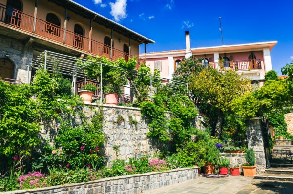 Panagia Kernitsis Monastery features lush gardens with vibrant flowers and stone walls under a bright blue sky in Arcadia, Greece.