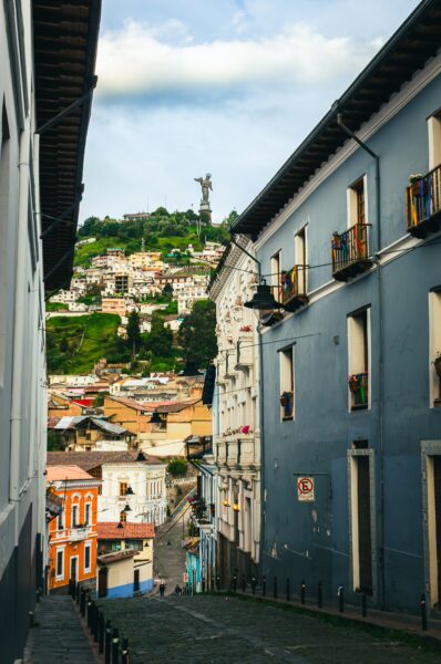 Colorful buildings line the cobblestone street, with the towering statue of the Virgin of El Panecillo visible in the background in Quito, Ecuador.