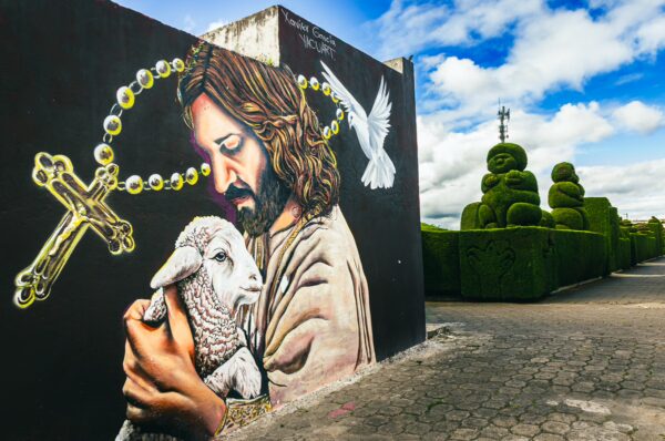 Vibrant mural depicting a Christ-like figure holding a lamb, surrounded by lush topiary at Tulcán Cemetery, Ecuador.