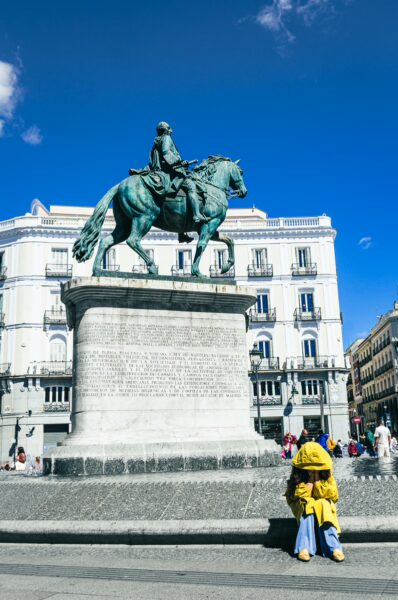Equestrian statue of Carlos III stands proudly in Madrid, with a vibrant blue sky and a person in a yellow raincoat seated nearby.