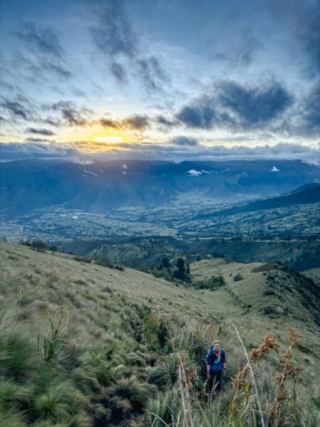 Stunning sunset over Imbabura Volcano, with a hiker navigating grassy slopes and distant valleys in Ecuador’s landscape.