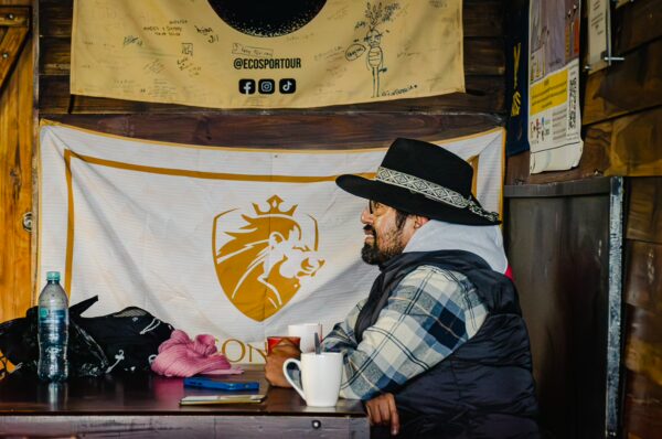 A man wearing a hat sits at a table inside the José Rivas Refuge, holding a mug with a flag in the background, Mount Cotopaxi, Ecuador.