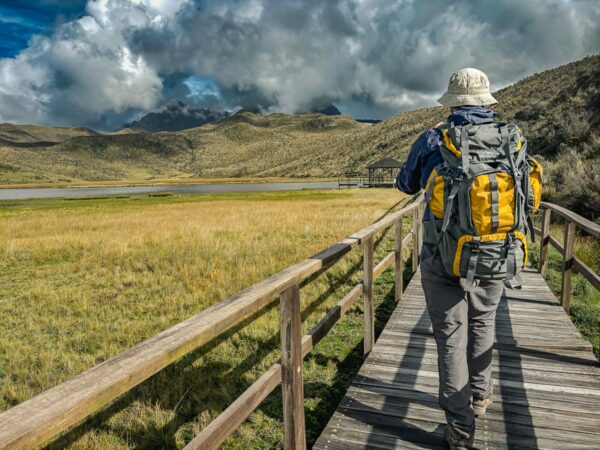 Hiker with a yellow backpack walking along a wooden path, with lush green grass and dramatic clouds over Laguna de Limpiopungo in Cotopaxi National Park.