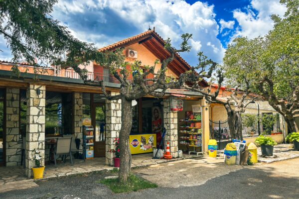 Quaint storefront with colorful displays under a blue sky in Nymfasia, Arcadia, Greece, surrounded by lush greenery.