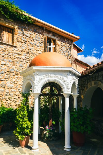 Panagia Kernitsis Monastery features a white colonnade structure with a red dome, surrounded by lush greenery and stone buildings in Arcadia, Greece.