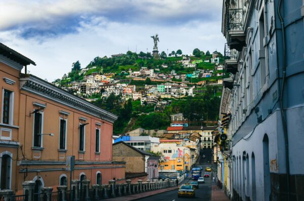 Colorful houses line the street, leading to the towering statue on a hillside in Quito, Ecuador, under a cloudy sky.
