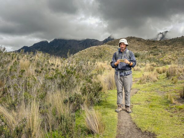 Hiker on a grassy trail with tall plants, surrounded by the dramatic landscape of Raminahui, Ecuador, under cloudy skies.