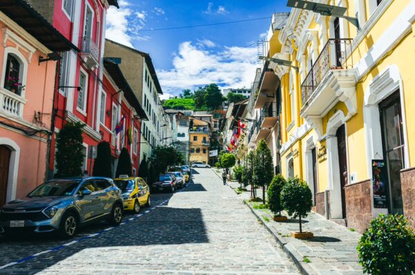 Colorful colonial buildings line the cobblestone street in Quito, Ecuador, under a bright blue sky dotted with clouds.