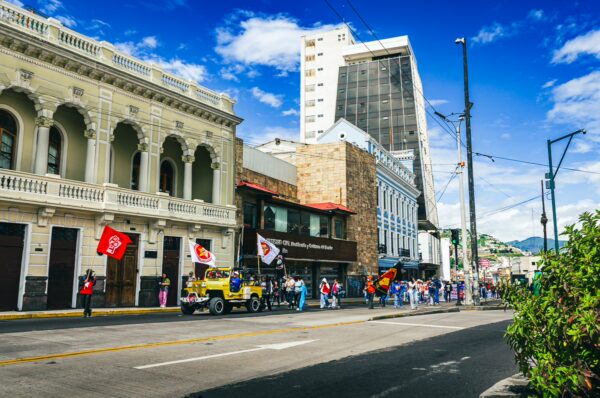 Colorful parade participants with flags march along a bustling street in Quito, Ecuador, framed by historic and modern buildings.
