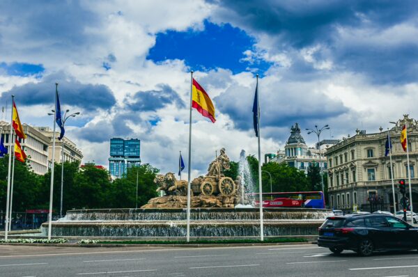 Cibeles Fountain in Madrid, surrounded by flags and lush greenery, under a dramatic sky with soft clouds.