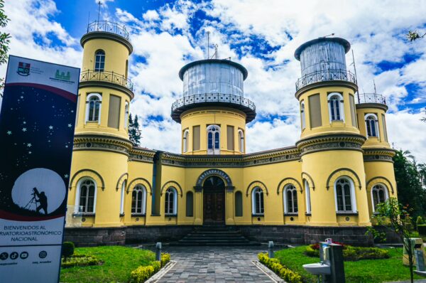 Yellow observatory with distinct towers, surrounded by vibrant greenery and a bright blue sky in Quito, Ecuador.