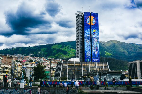 Vibrant street art decorates a modern building in Parque La Alameda, with lush green hills and cloudy skies framing Quito, Ecuador.