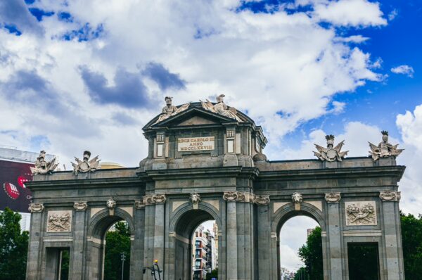 Puerta de Alcalá stands majestically under a blue sky with clouds, showcasing intricate stone carvings and green trees in Madrid.