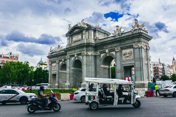Puerta de Alcalá stands majestically in Madrid, with clouds overhead and a bustling street scene featuring vehicles and greenery.
