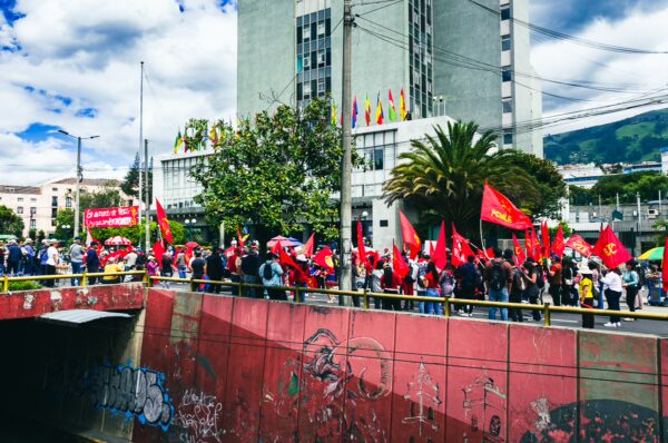 Red flags wave in a vibrant parade in Quito, Ecuador, showing diverse crowds celebrating International Workers' Day against a backdrop of modern buildings.