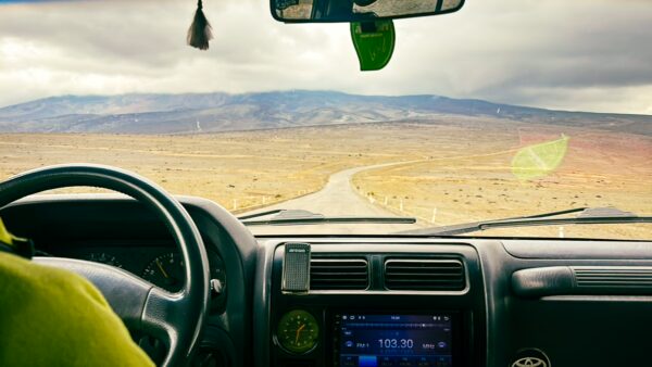A car's interior view shows an open road leading toward the majestic Chimborazo mountain in Ecuador under a cloudy sky.