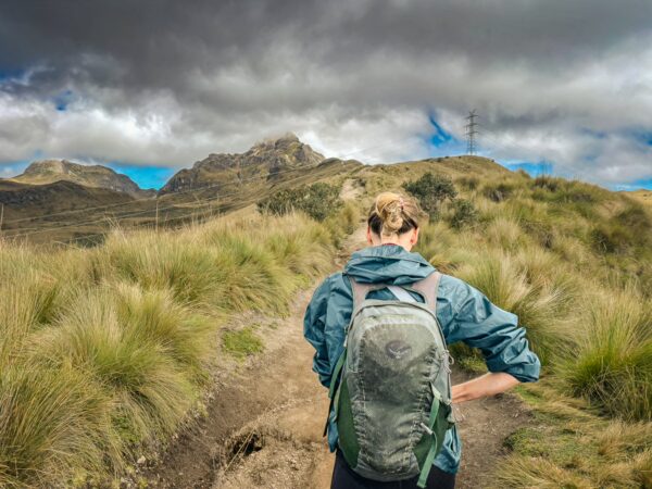 Hiker with a backpack walking through grassy terrain under dramatic clouds, with Pichincha Volcano looming in the background, Ecuador.