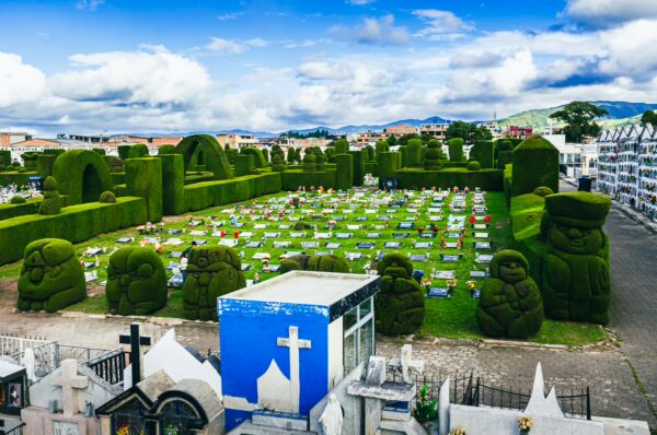 Green manicured hedges and unique sculptures create a striking landscape at Tulcán Cemetery in Ecuador, with white graves punctuating the scene.