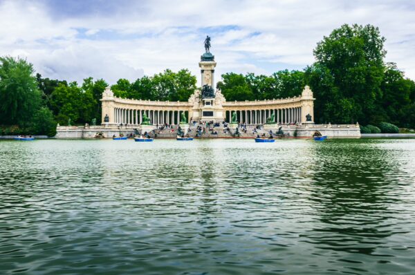 Boats glide across the tranquil water in Parque El Retiro, featuring a grand monument surrounded by lush greenery in Madrid, Spain.