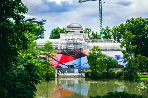 Colorful art wraps the Palacio de Cristal in Parque El Retiro, Madrid, surrounded by lush greenery and a tranquil pond.