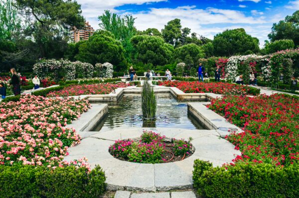 Colorful flowers and lush greenery surround a tranquil fountain at Parque El Retiro, Madrid, Spain, creating a serene atmosphere.