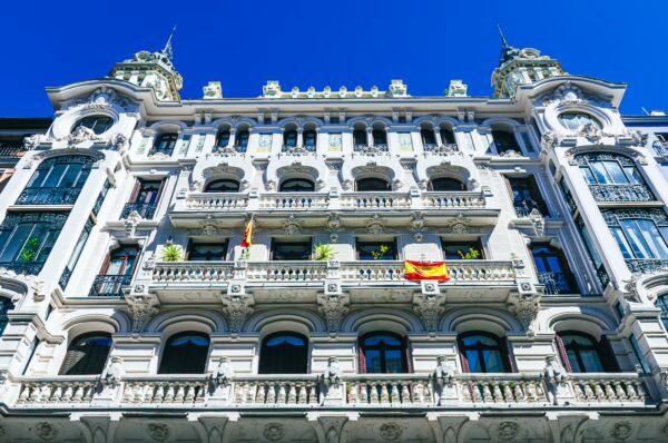 Elegant, historic building with ornate balconies and Spanish flags, set against a vibrant blue sky in Madrid, Spain.