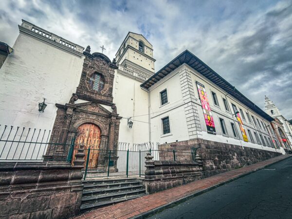 Historic architecture lines a street in Quito, Ecuador, featuring a grand stone entrance and detailed facades under a moody sky.