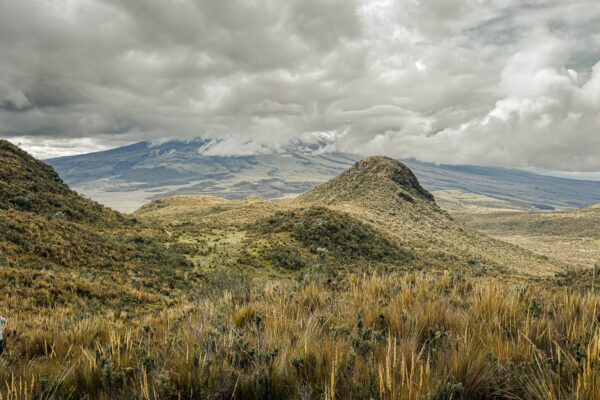 Volcan Ruminahui rises in the background, surrounded by rolling hills and lush green vegetation under a dramatic cloudy sky in Ecuador.