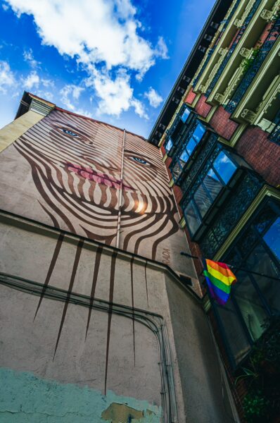Vibrant mural featuring a face on a building wall with a rainbow flag hanging beside it in Madrid, Spain. Bright blue sky enhances the scene.