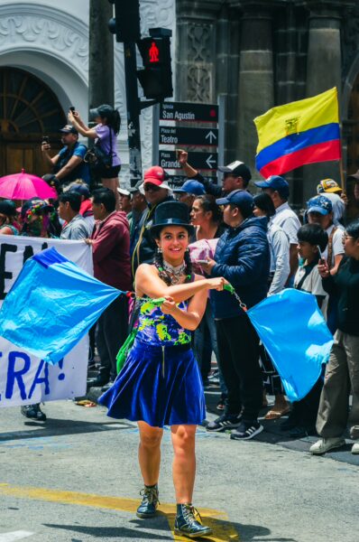 Colorfully dressed dancer waves blue flags at the International Workers' Day parade in Quito, Ecuador, surrounded by festive crowds.