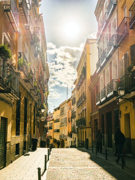 Charming sunlit street in Madrid, Spain, lined with colorful buildings, balconies, and cobblestone pavement under a bright sky.