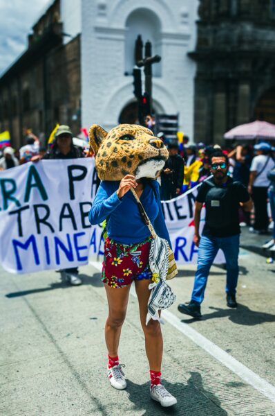Festive parade for International Workers' Day in Quito, Ecuador, featuring a participant in a leopard mask and colorful attire.