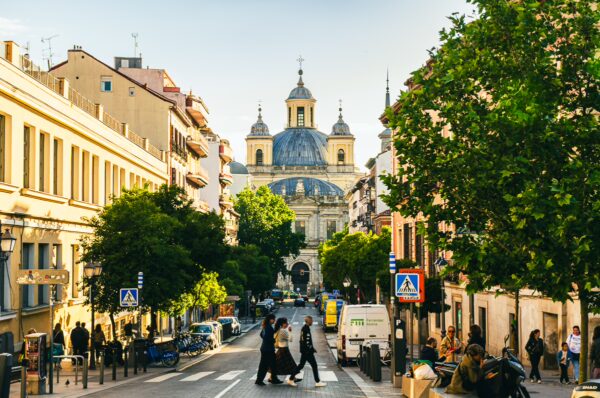 Bustling street scene in Madrid features tree-lined avenues and the stunning dome of a historic building in the background.